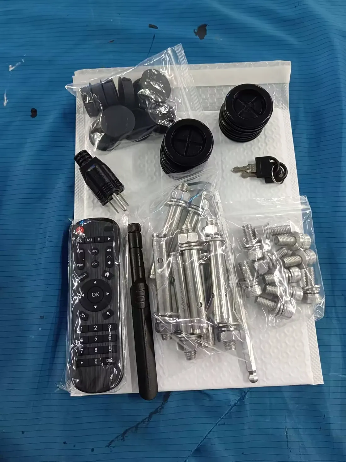 The supplied accessory kit laid out on a bench: a remote control, a stubby Wi-Fi antenna, a power plug, two keys on a ring, black plastic caps and feet, eight large M12 expansion bolts, a bag of smaller bracket screws and a hex key.