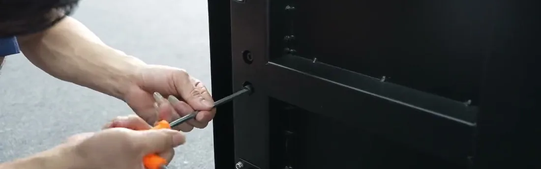 Close-up of two hands driving a screw with a hex driver through the vertical bracket rail into the back of the display cabinet, which is lying flat.