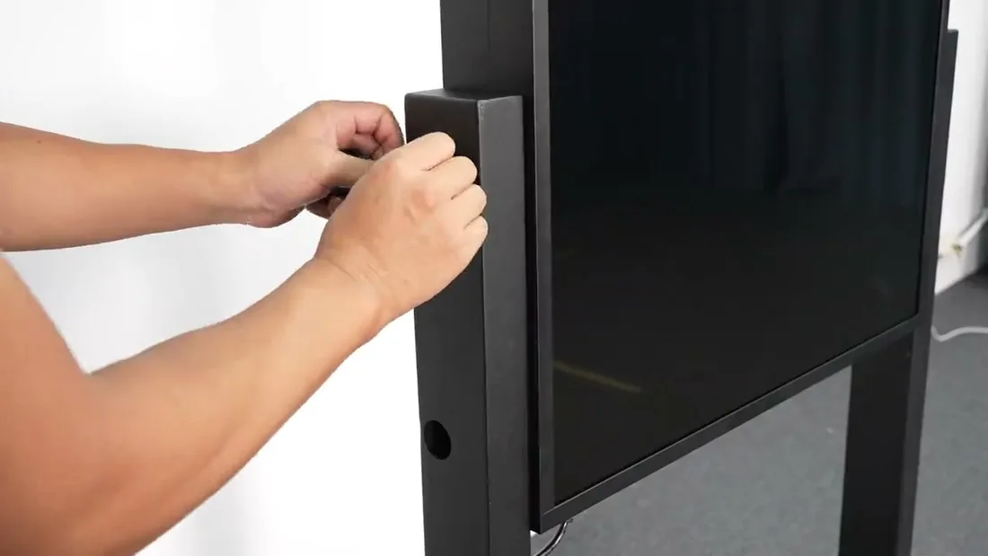 Two hands gripping the vertical bracket rail of the assembled display while it stands upright, with the black screen visible alongside and nobody touching the glass.